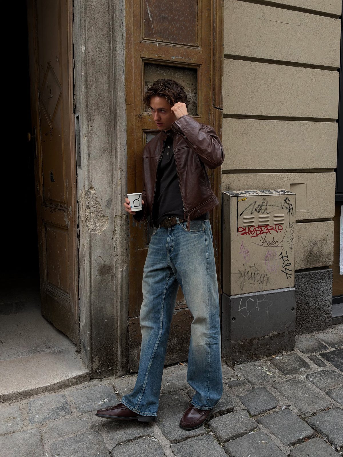 Man in brown leather jacket and blue jeans standing on a cobblestone street