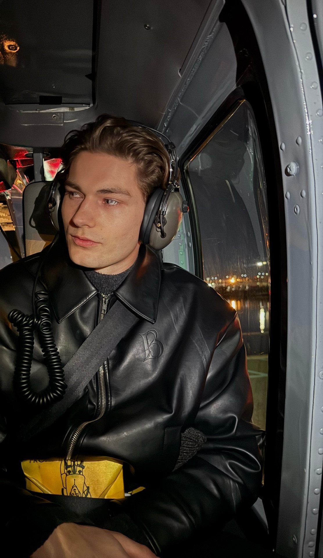 Man in a black leather flight jacket wearing aviation headsets inside a helicopter.