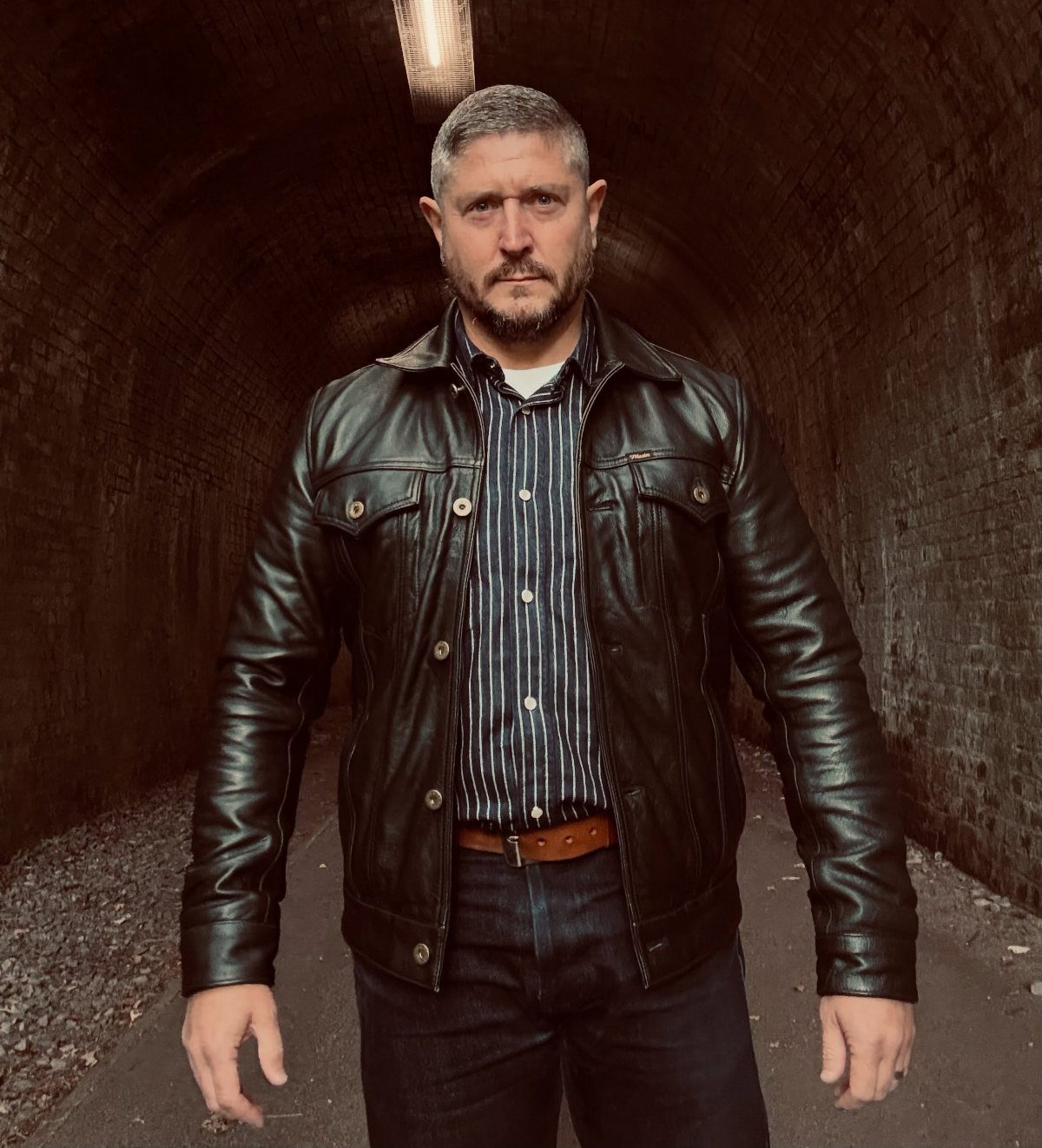 Man wearing a black leather jacket and striped shirt in a brick tunnel