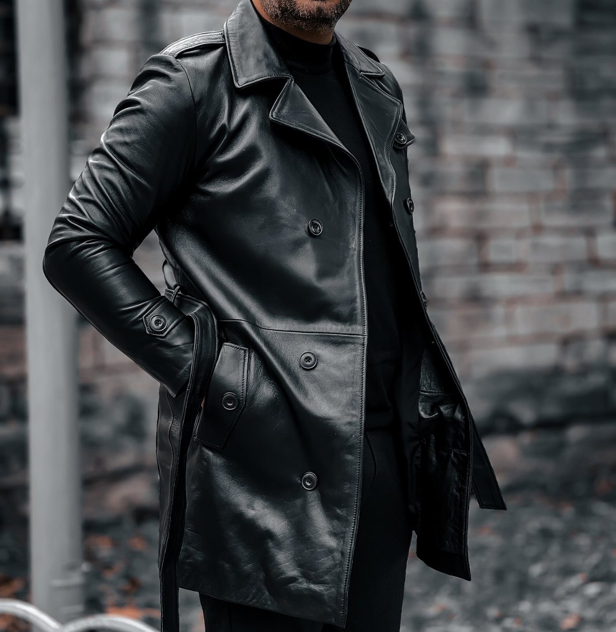 Man wearing a long black leather car coat and black turtleneck against brick wall