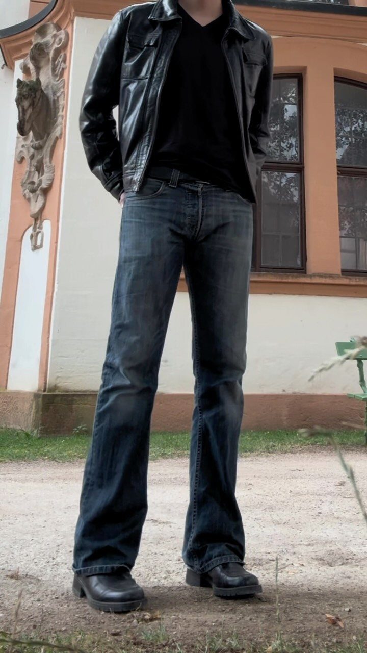 Person wearing a black leather jacket and flared blue jeans standing outdoors.