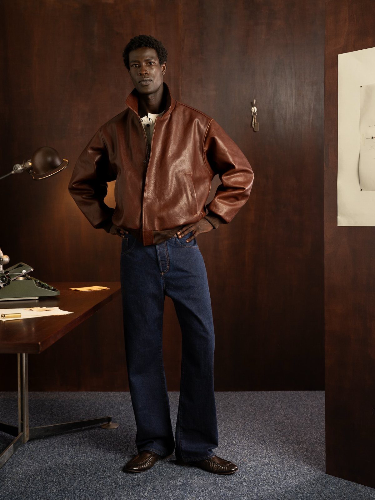 Man wearing a brown leather jacket and wide-leg blue jeans in a wood-paneled room
