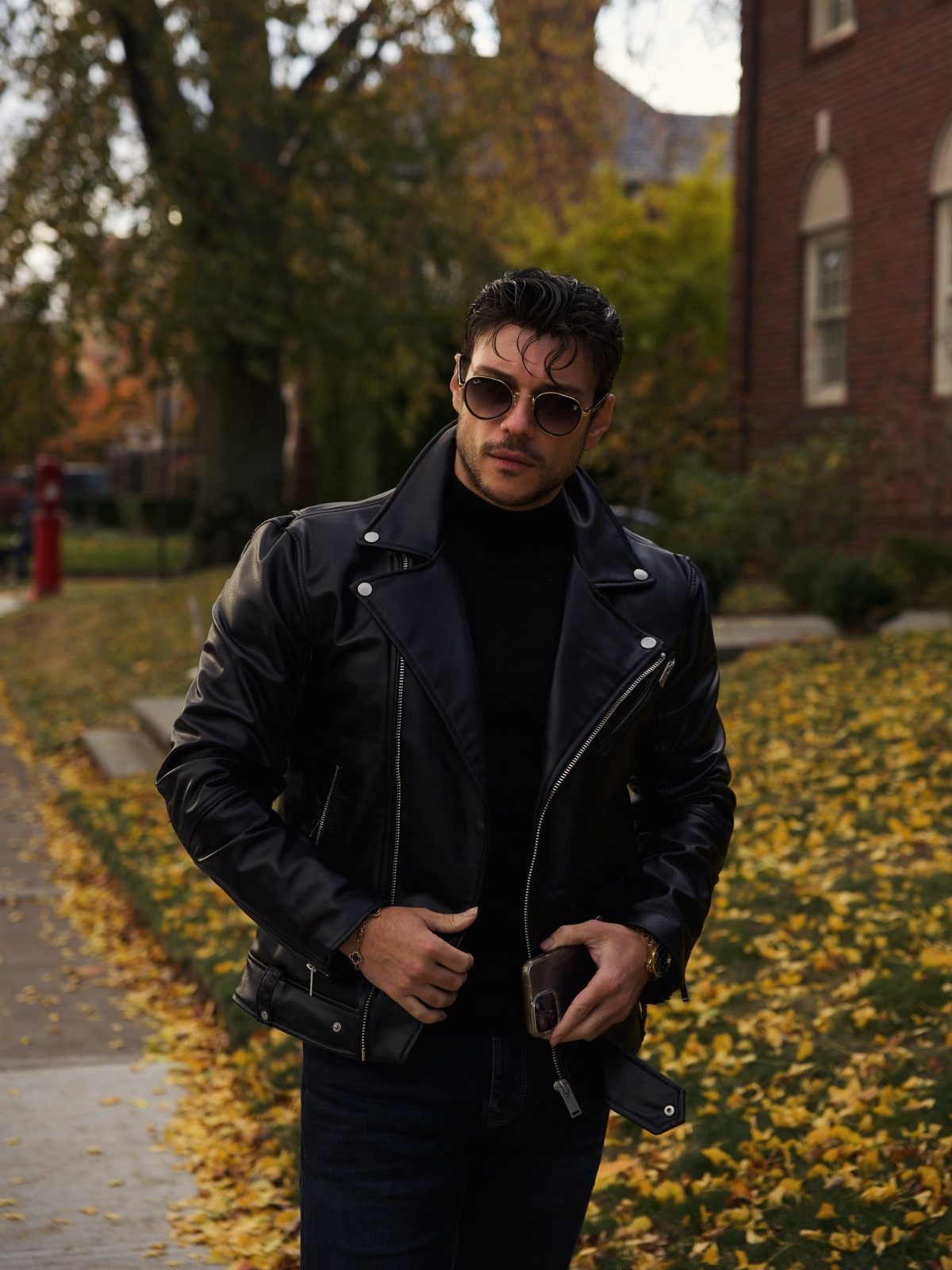 Man in black leather biker jacket and turtleneck standing among autumn leaves.