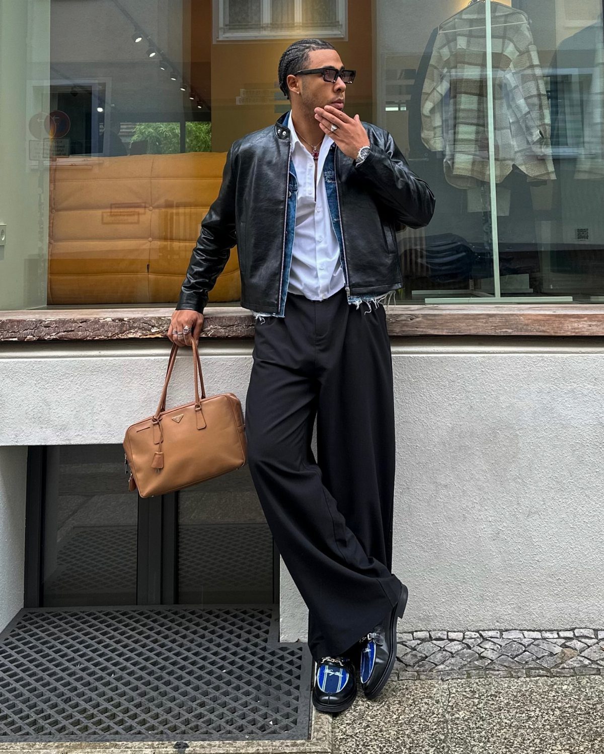 Man in black leather jacket, denim shirt, and wide black trousers holding tan bag.