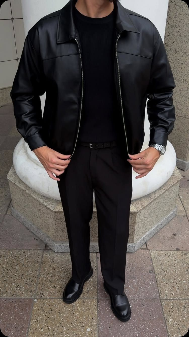 Man wearing a black leather jacket, black t-shirt, and black trousers standing outdoors.