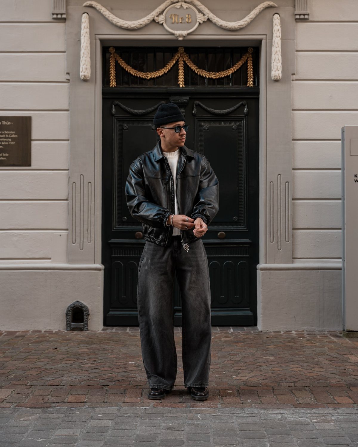 Man in black leather jacket and wide grey trousers standing before ornate door