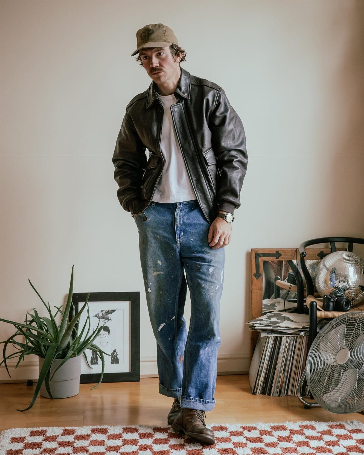 Man wearing a brown leather flight jacket, white tee, and blue jeans indoors.