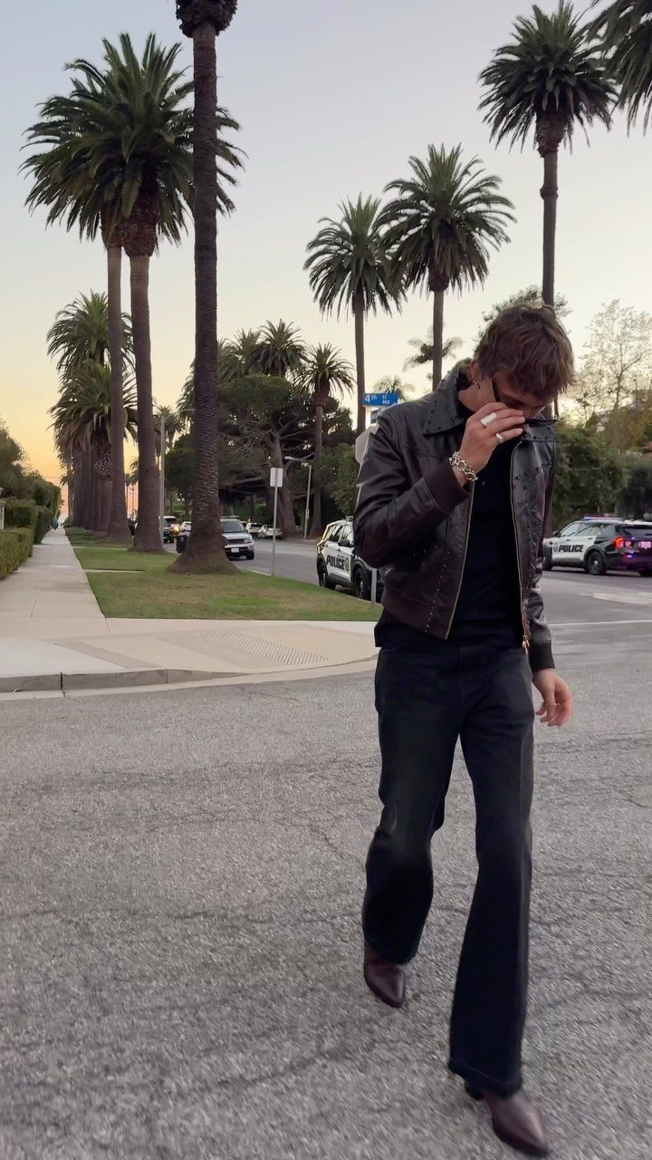 Man wearing a brown leather jacket and brown leather boots on a palm-lined street