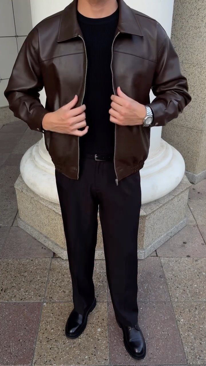 Man wearing a brown leather jacket and black trousers standing by a white column