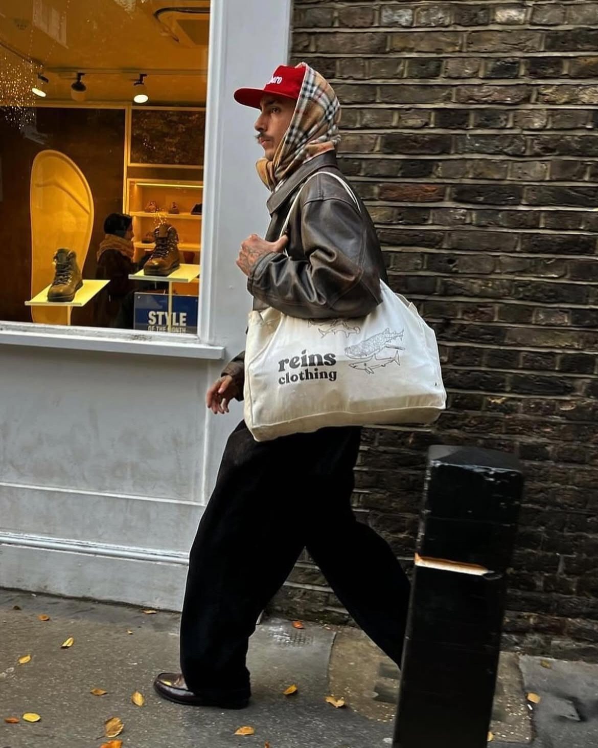 Person in brown leather jacket, plaid scarf, and black trousers walking on sidewalk