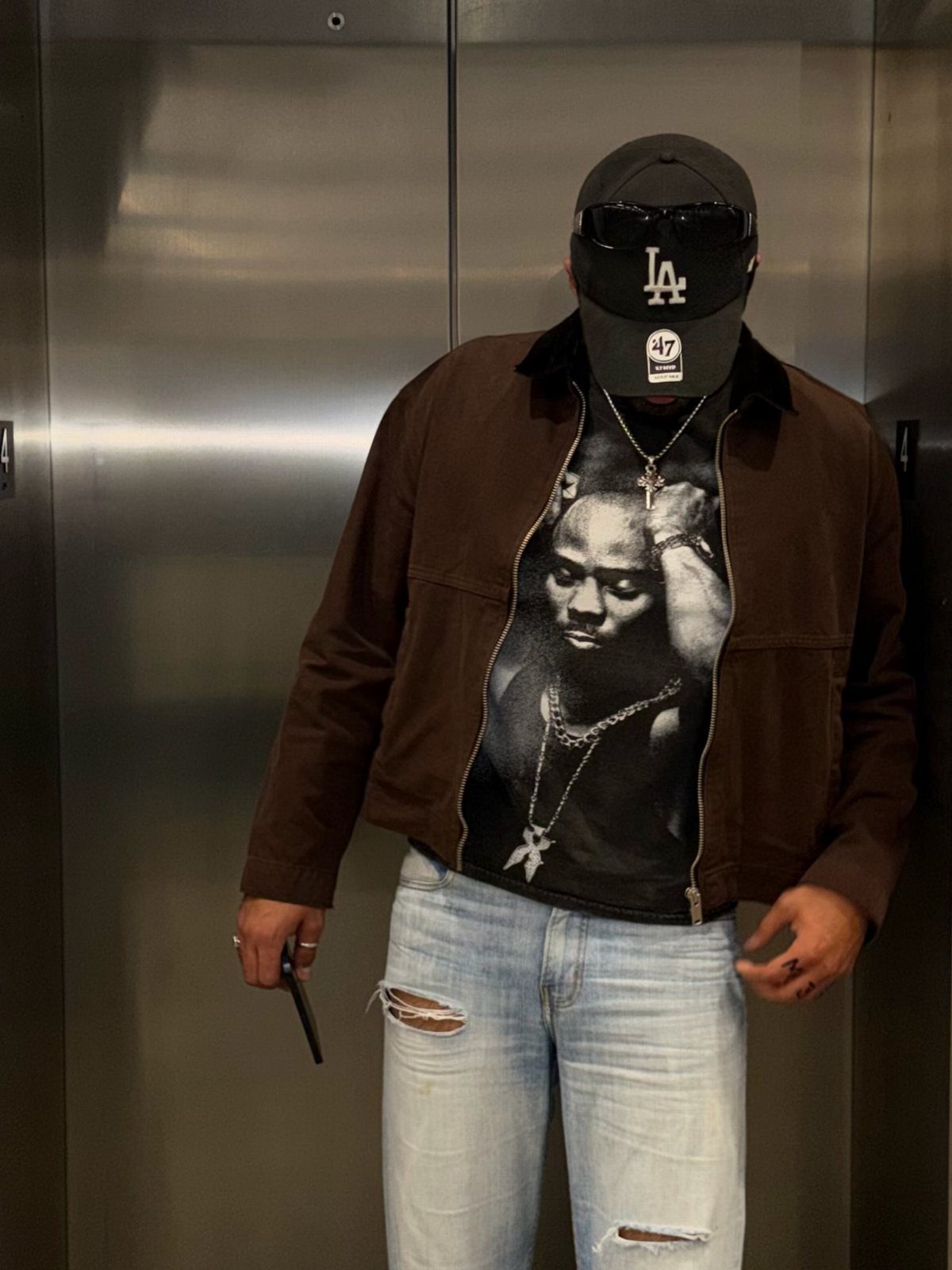Person in brown jacket and graphic tee with ripped light blue jeans and cap.