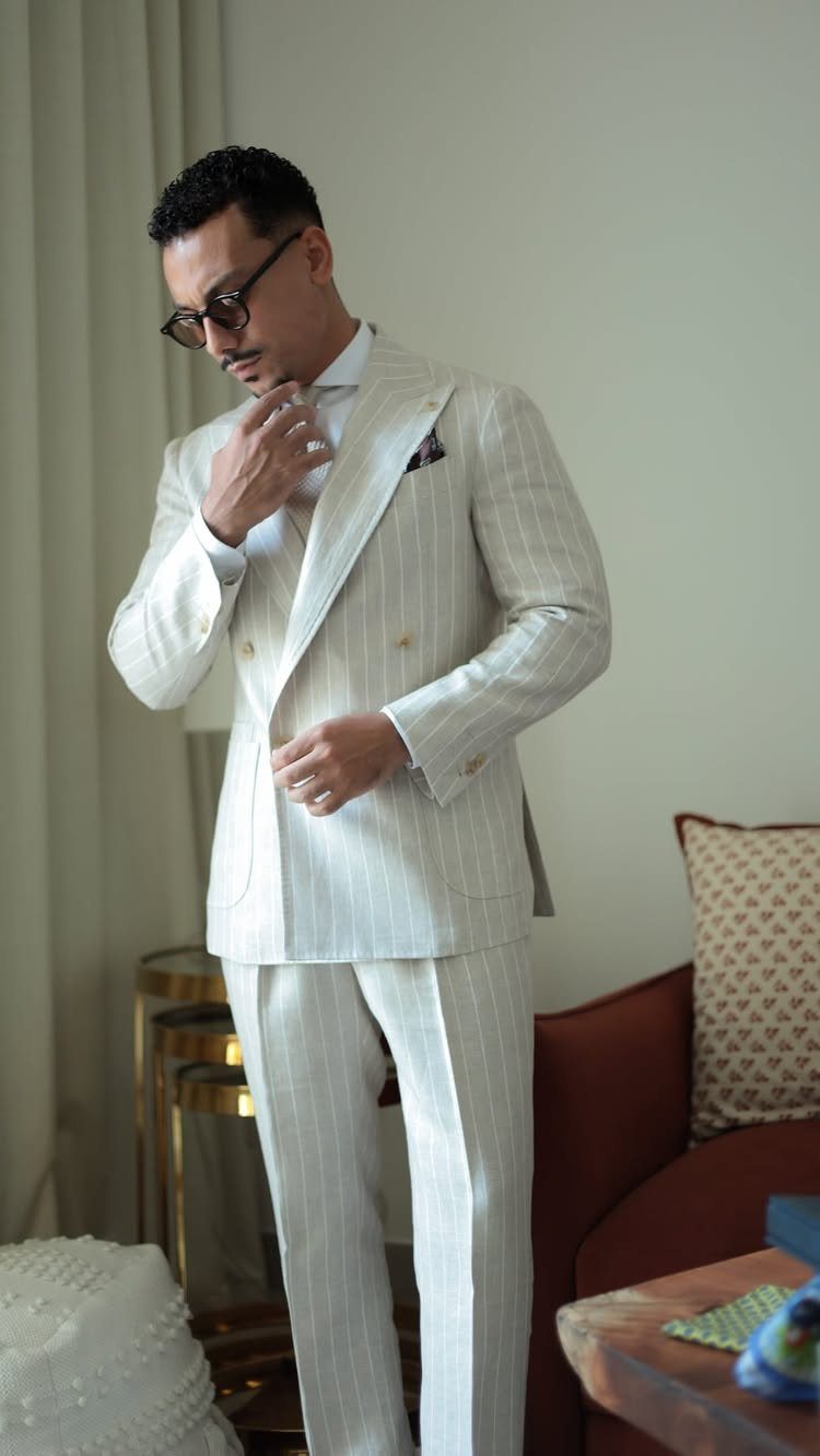 Man in a light pinstriped linen suit adjusting his tie indoors.