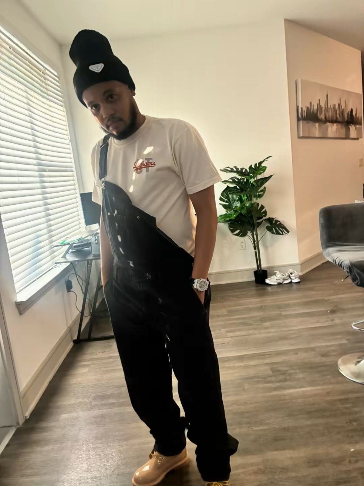 Man wearing black bib overalls, a white t-shirt, and tan boots indoors.