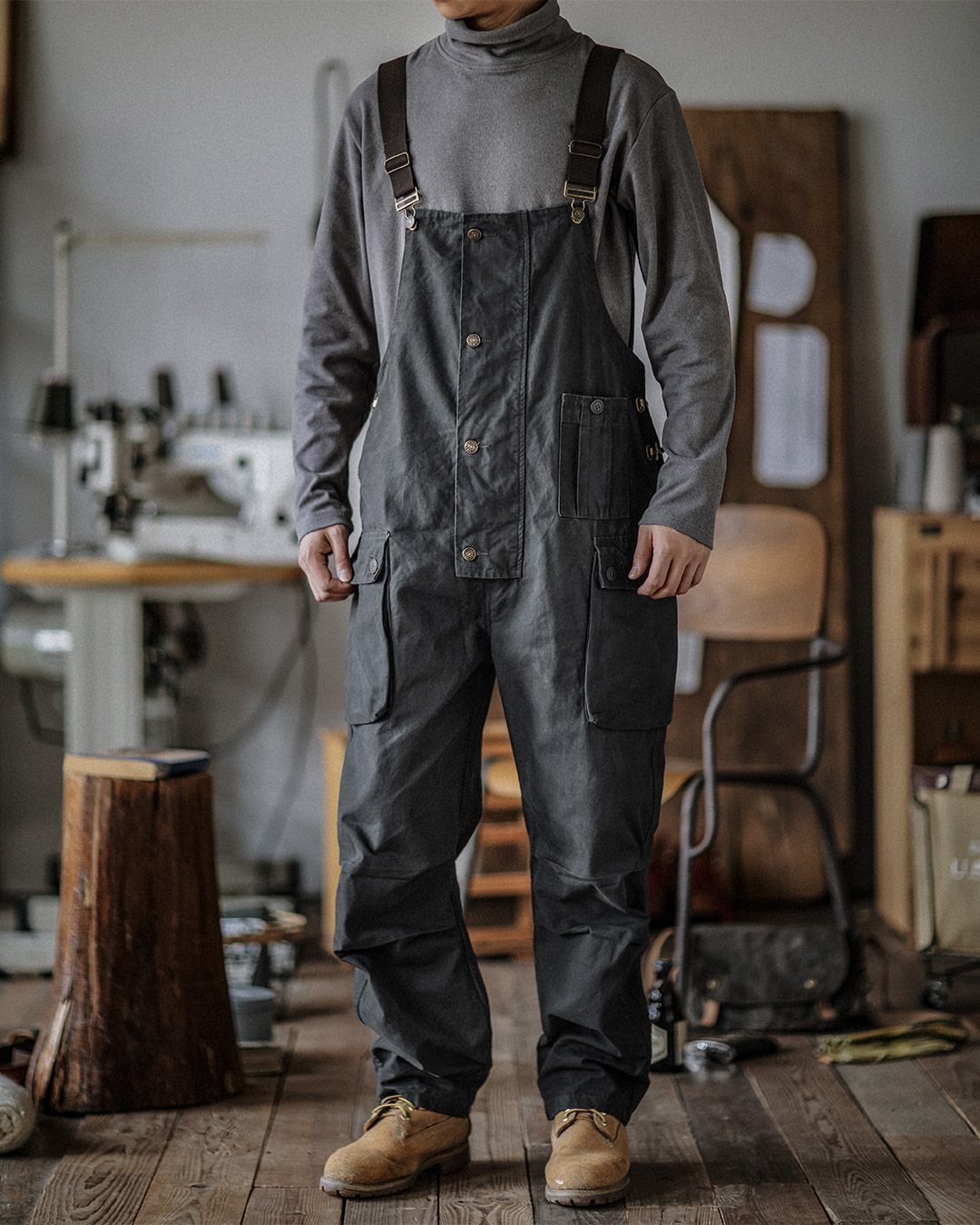 Man wearing black bib overalls and tan boots in a rustic workshop setting