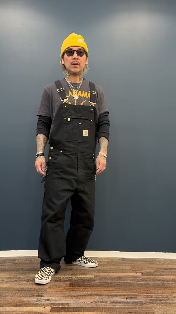 Man wearing black overalls, a yellow beanie, and checkered shoes against a blue wall.