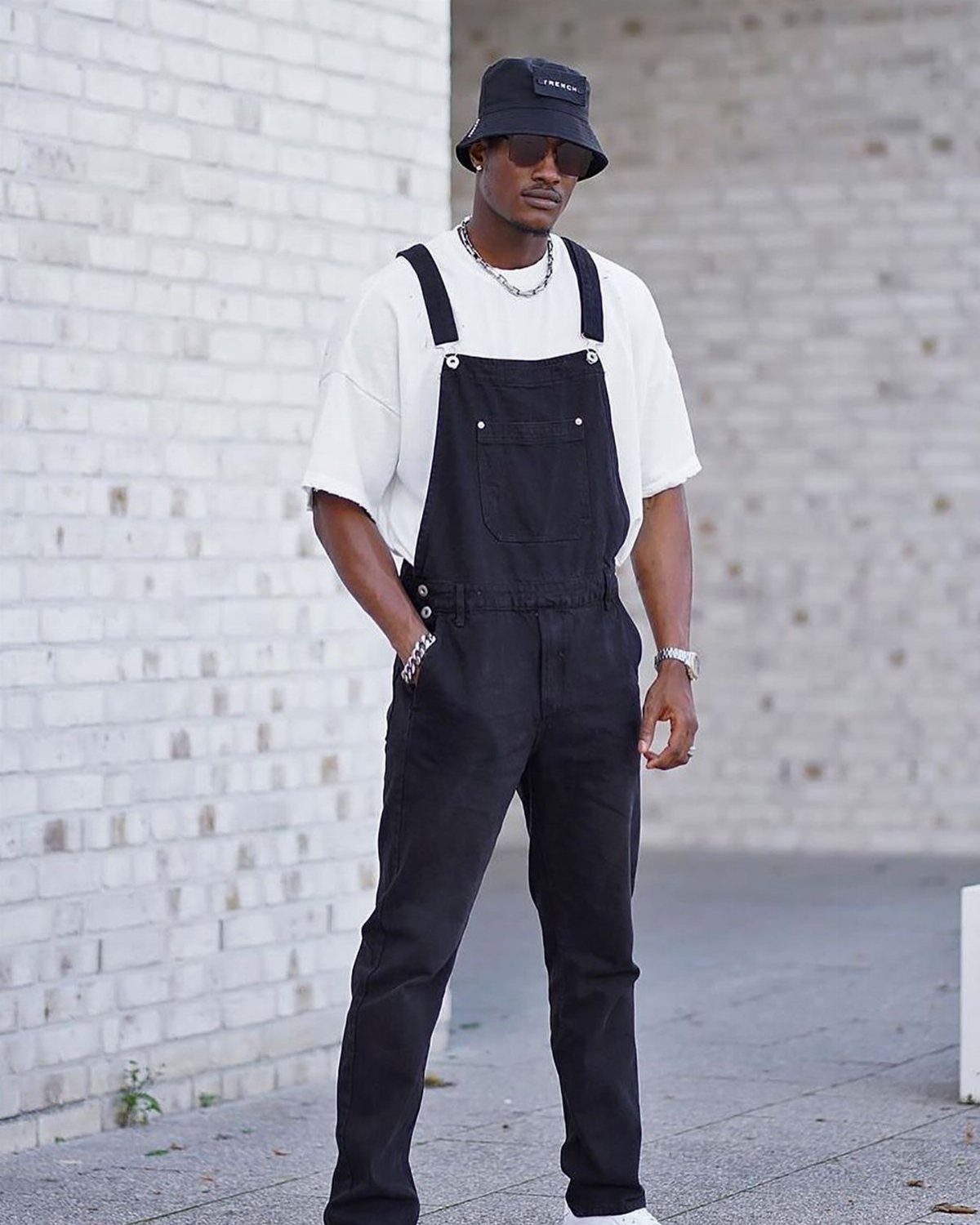 Man wearing black bib overalls and a white t-shirt against a brick wall