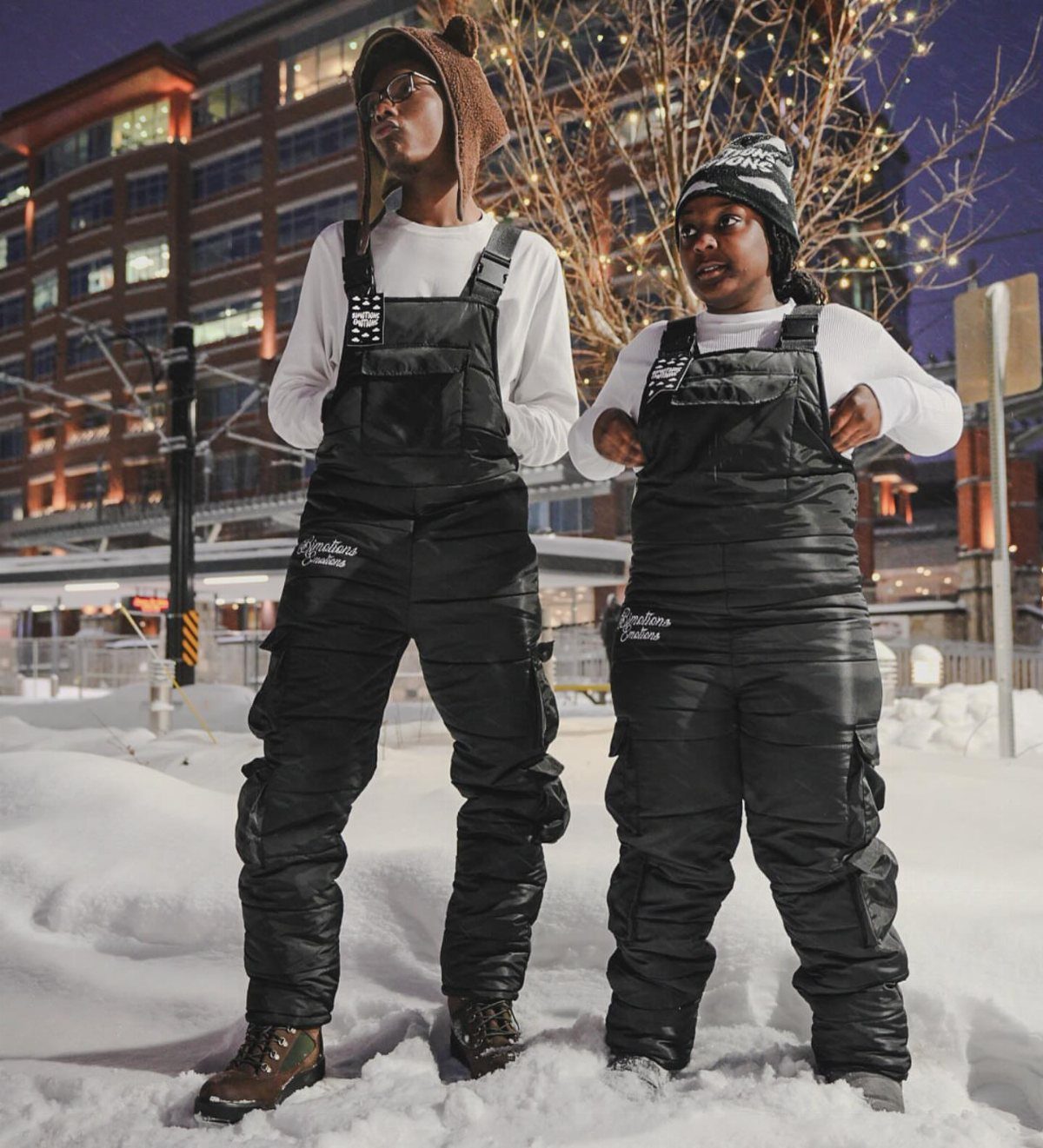 Two people wearing black bib overalls standing in a snowy outdoor urban setting.