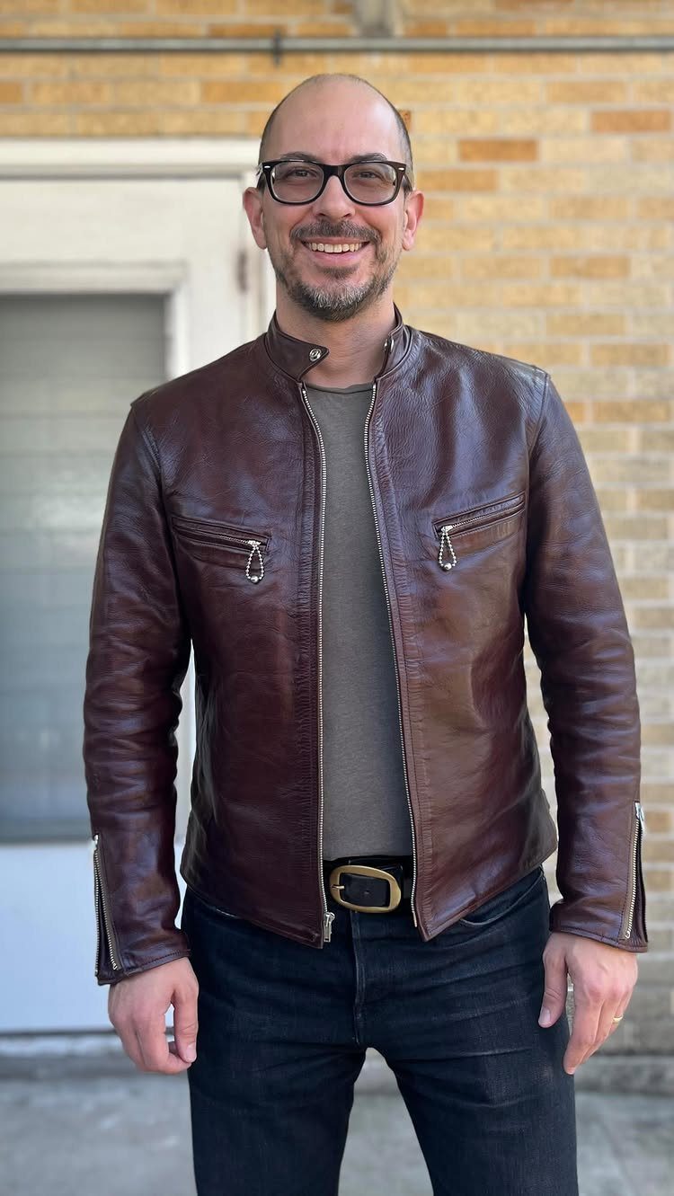 Man wearing a brown leather biker jacket and grey shirt with black jeans.