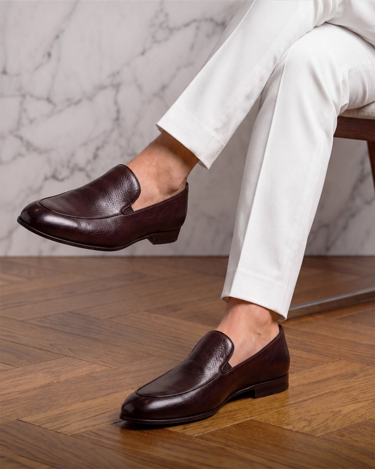 Dark burgundy leather loafers worn with white trousers on a wooden floor