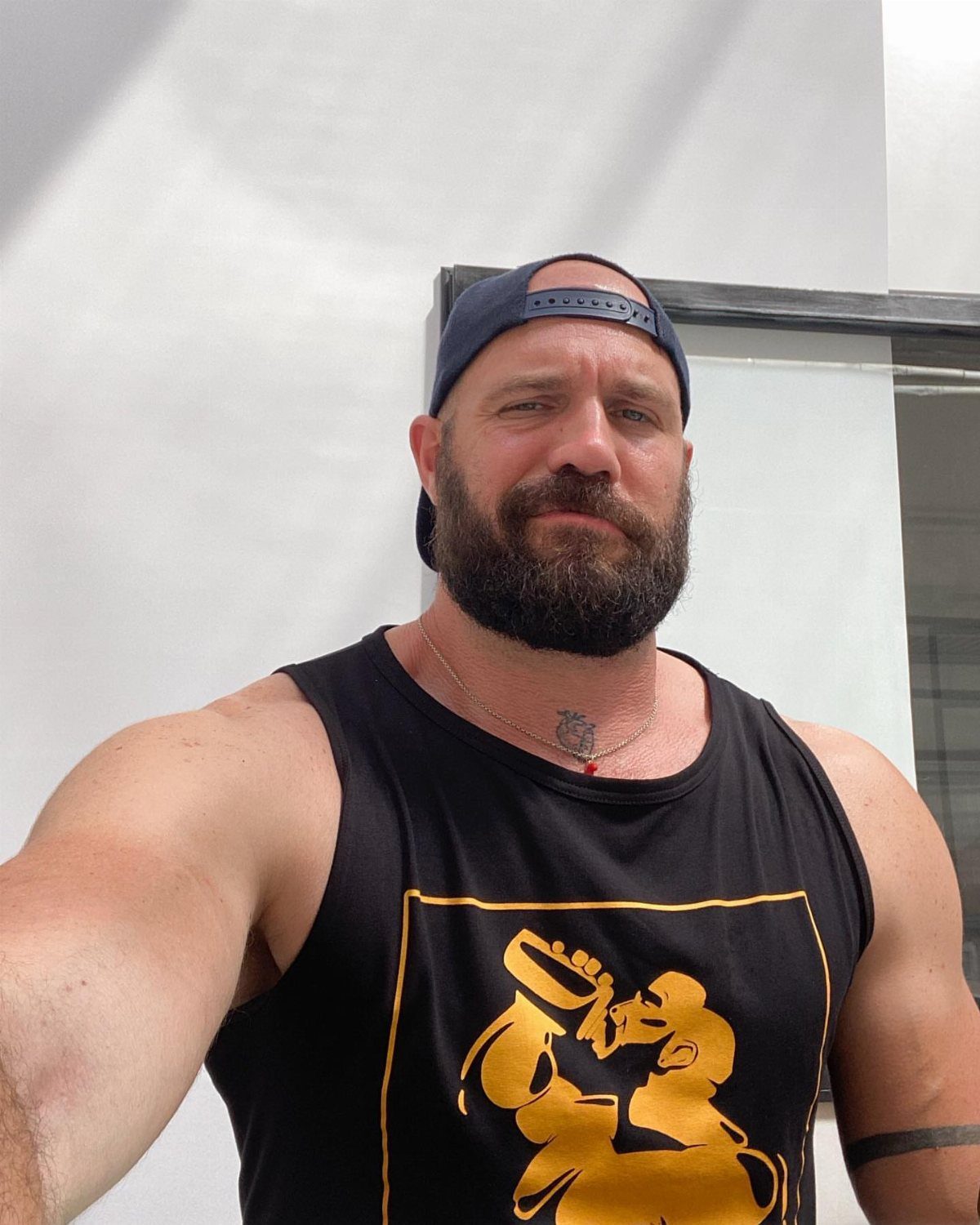 Bearded man wearing a black graphic tank top and blue baseball cap outdoors.