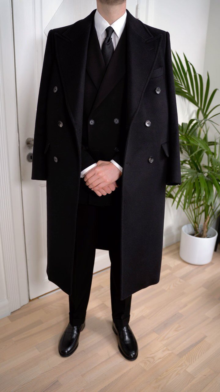 Man wearing a black overcoat over a black three-piece suit and black shoes.