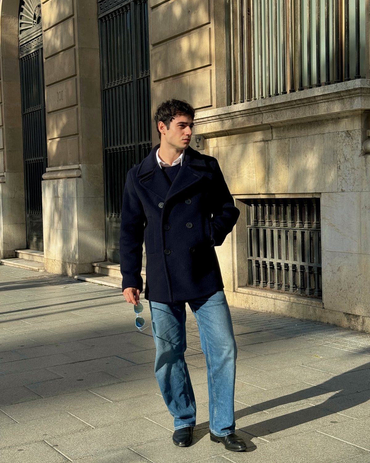 Man wearing a navy blue double-breasted peacoat and blue jeans standing outdoors.