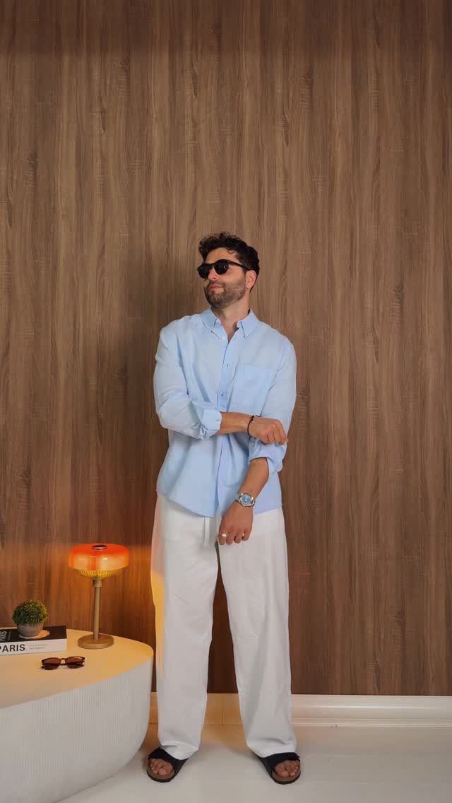 Man wearing a light blue shirt and wide white trousers against wood paneling.