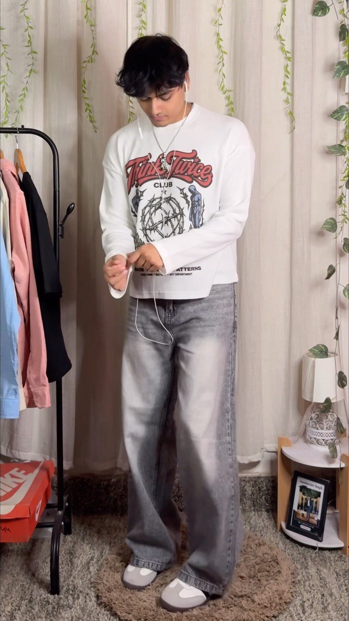 Young man wearing a white graphic long-sleeve shirt and wide-leg grey jeans.