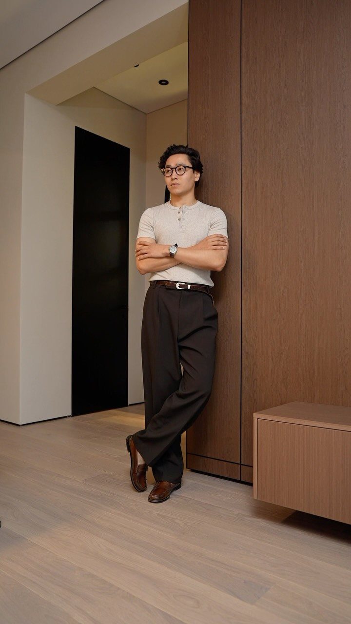Man in cream henley and dark brown trousers leaning against a wooden wall.