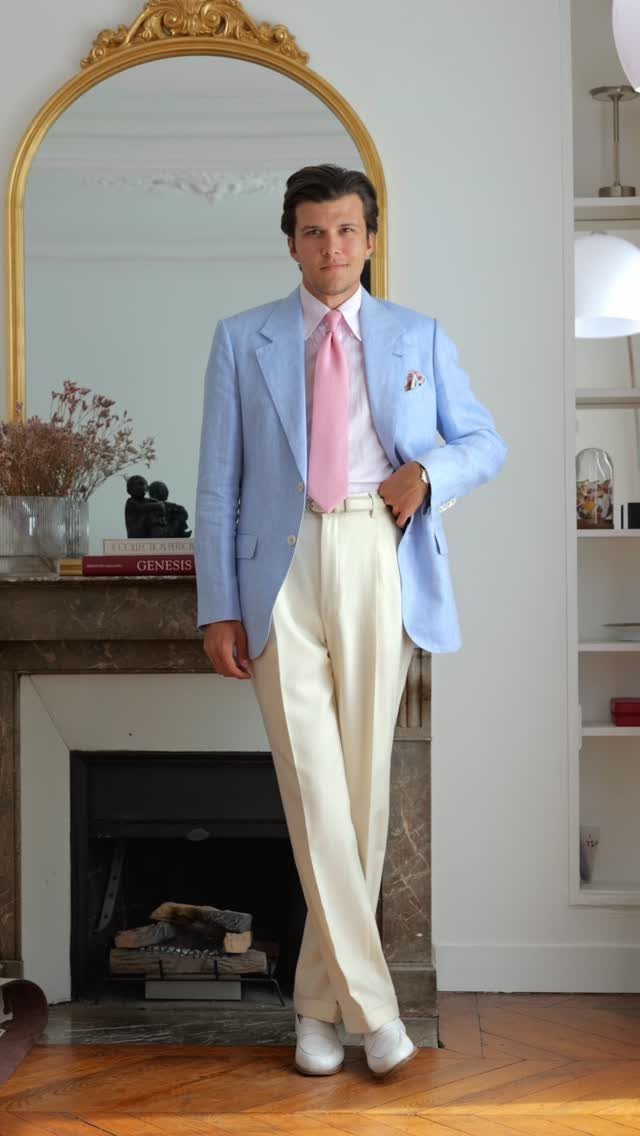 Man wearing a light blue blazer, pink tie, and cream trousers standing indoors.