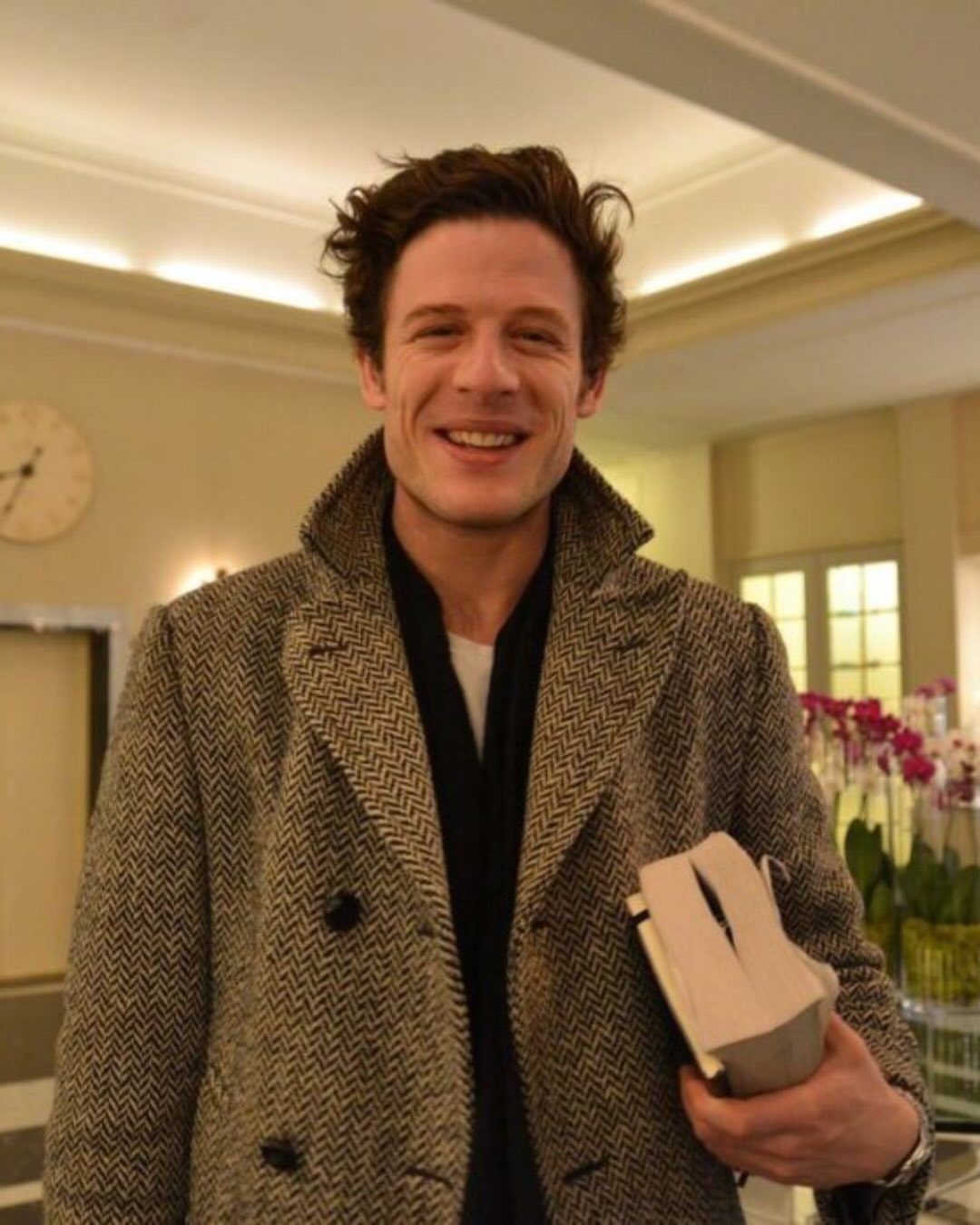 Man wearing a grey herringbone peacoat and black shirt holding a book indoors