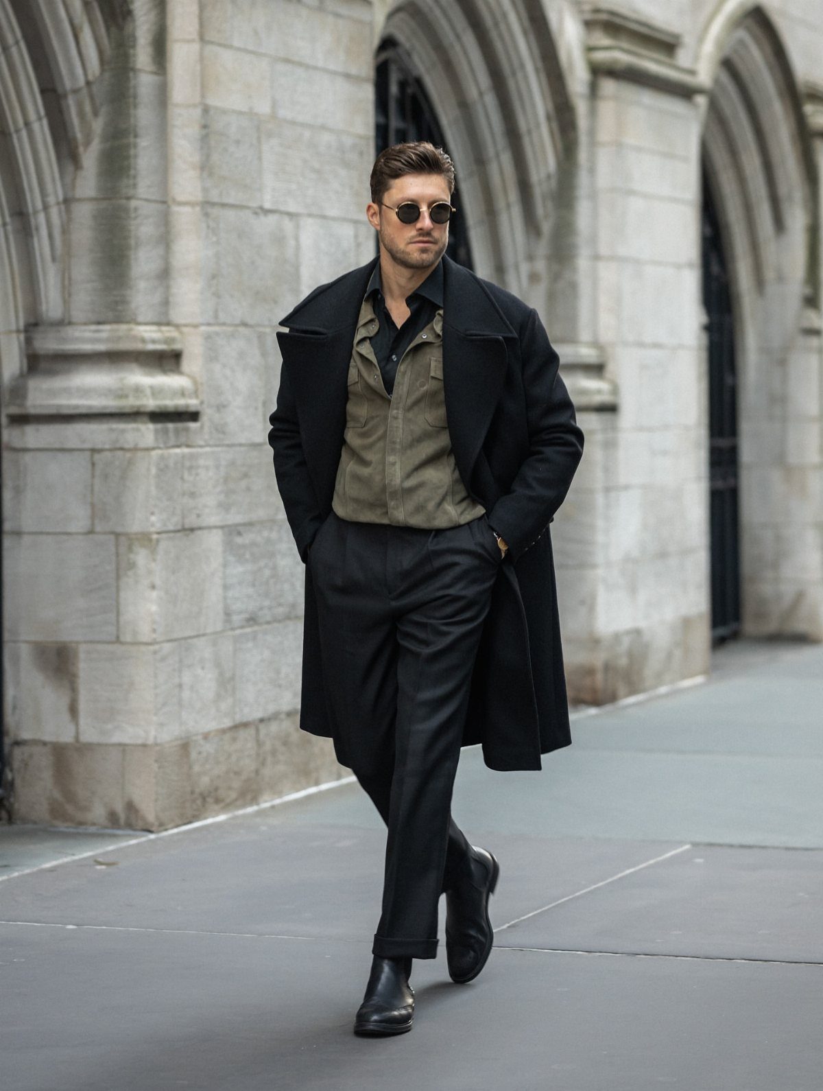 Man in a black peacoat, olive shirt, and black trousers standing outdoors.