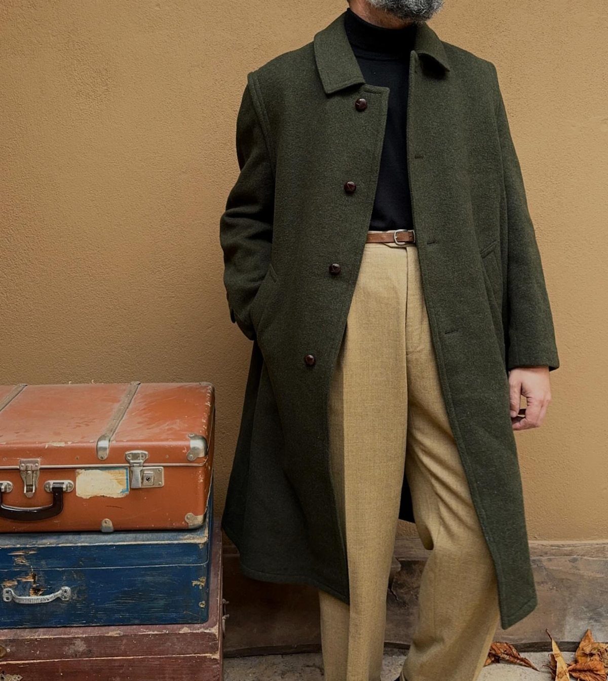 Man wearing a long dark green wool coat and tan trousers by vintage suitcases.