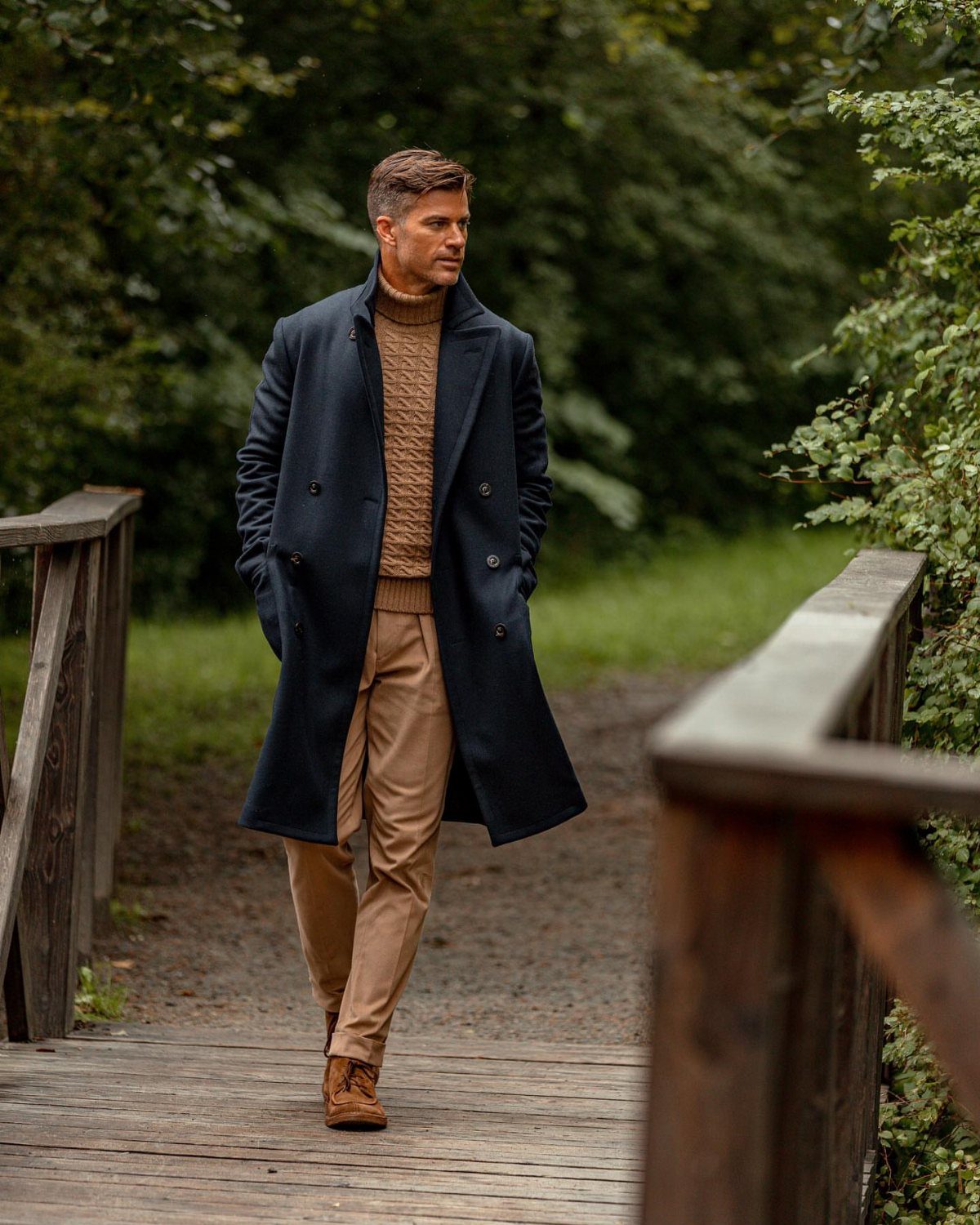 Man in navy blue wool coat and tan sweater walking on wooden bridge