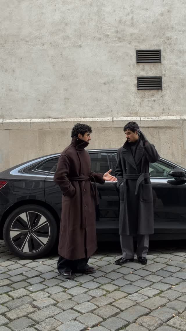 Two men wearing long wool wrap coats standing by a black car on cobblestones.