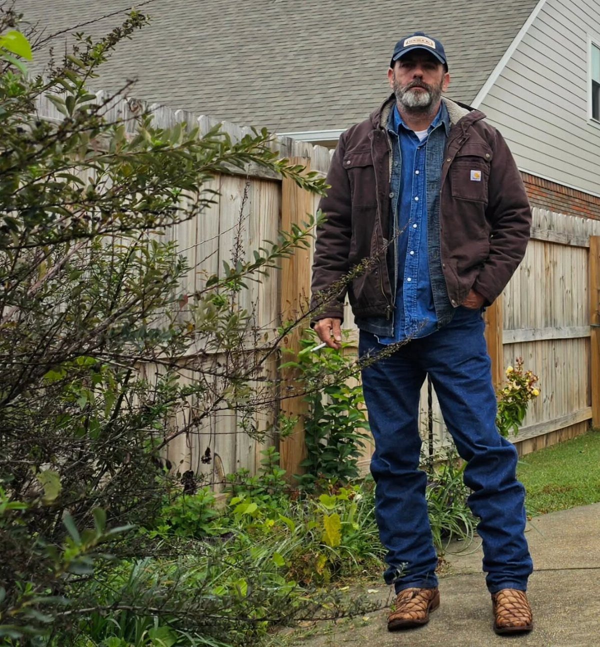 Man wearing a brown canvas field coat, denim shirt, and blue jeans outdoors.