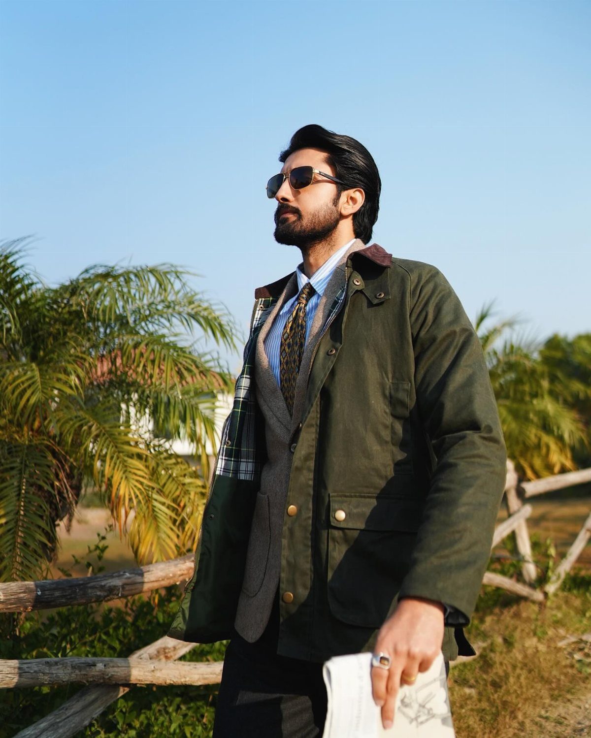 Man in a green field coat and sunglasses standing by a rustic wooden fence.