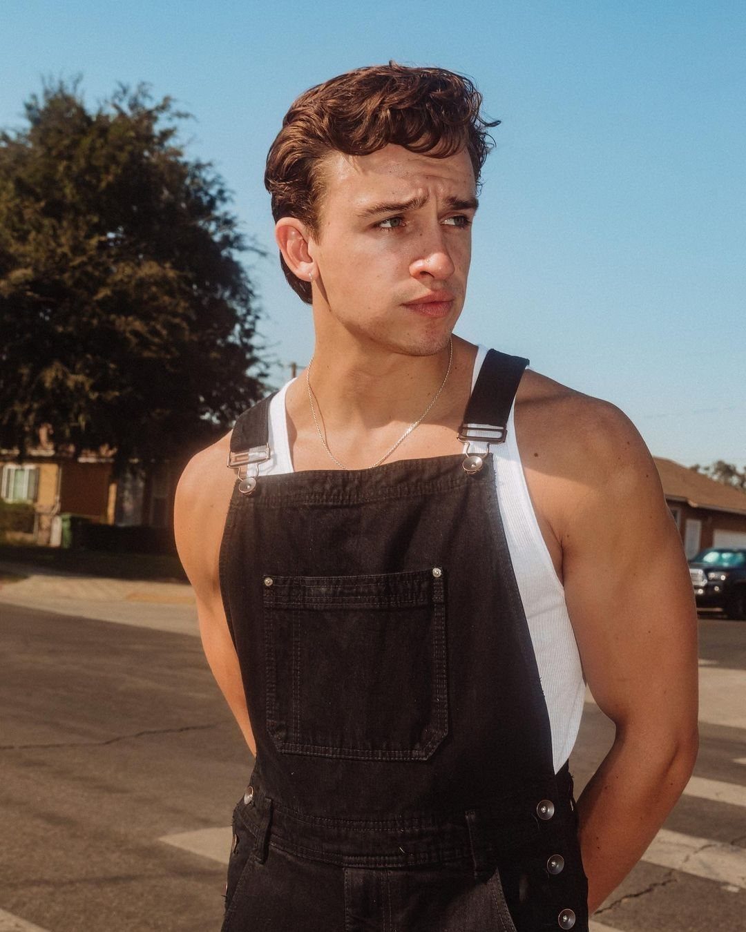Young man wearing black denim overalls and a white tank top outdoors.
