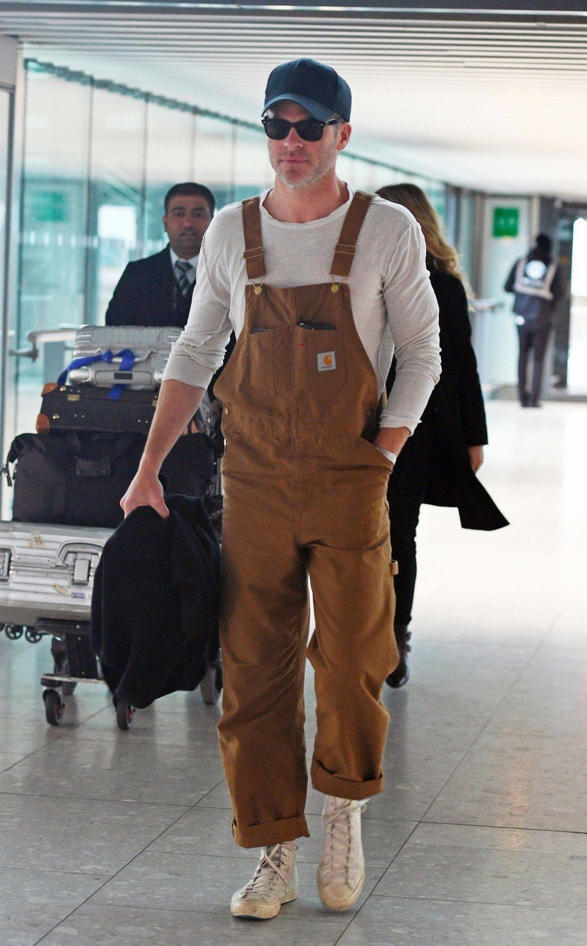 Man wearing brown canvas overalls and a baseball cap walking through an airport terminal