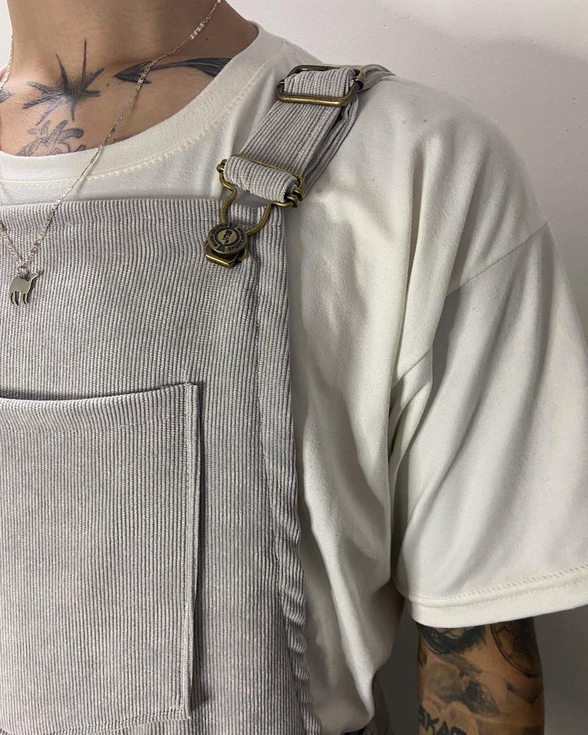 Light grey corduroy bib overalls worn over a white t-shirt with metal buckles.