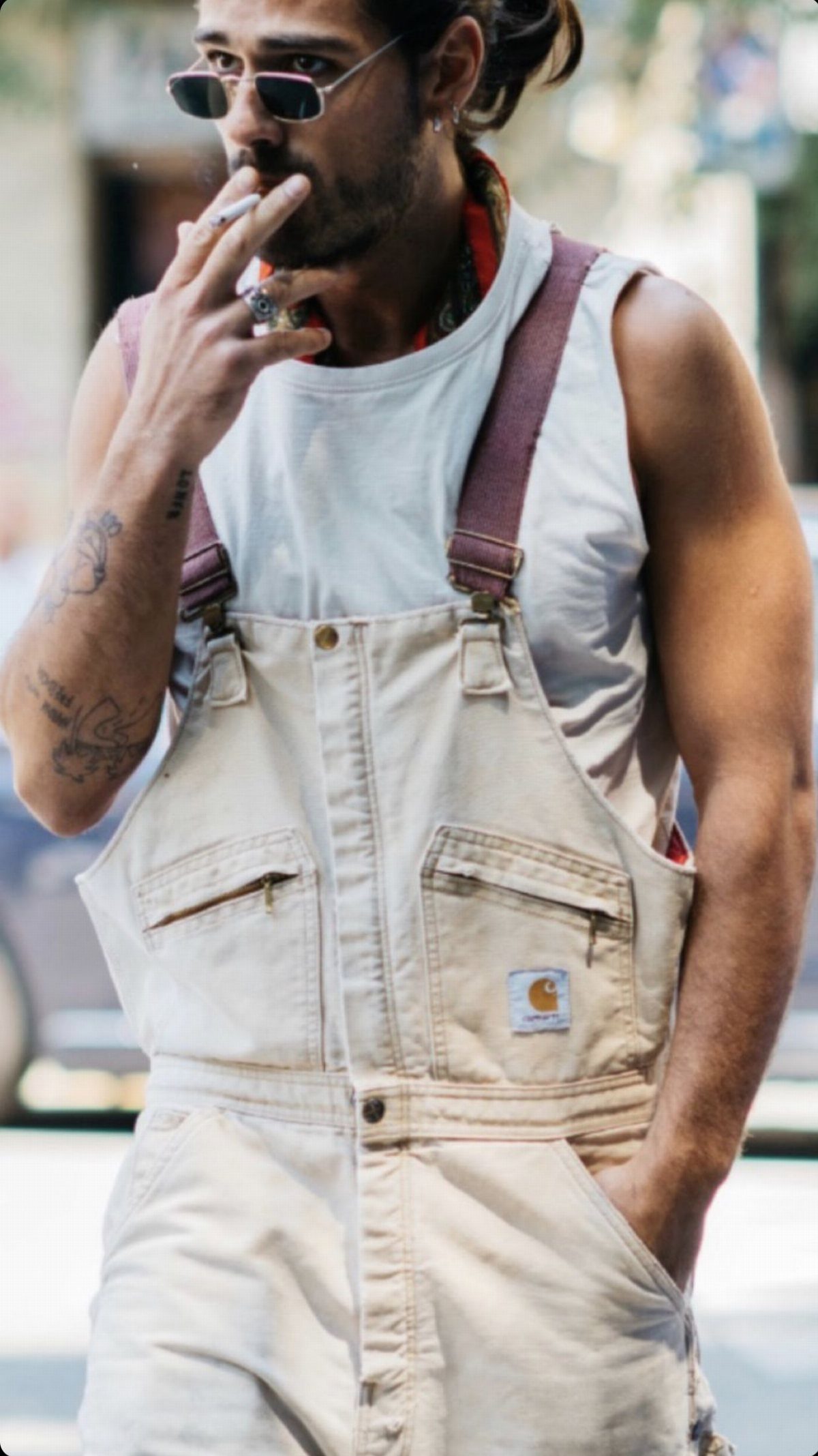 Man with tattoos wearing cream overalls and sunglasses smoking a cigarette outdoors.