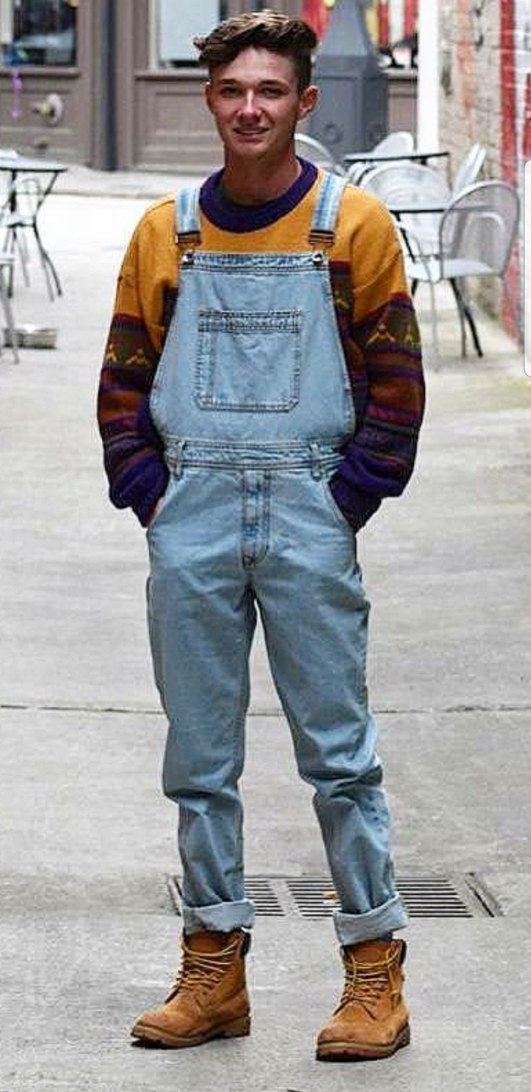 Young man wearing light blue denim bib overalls and a colorful patterned sweater.