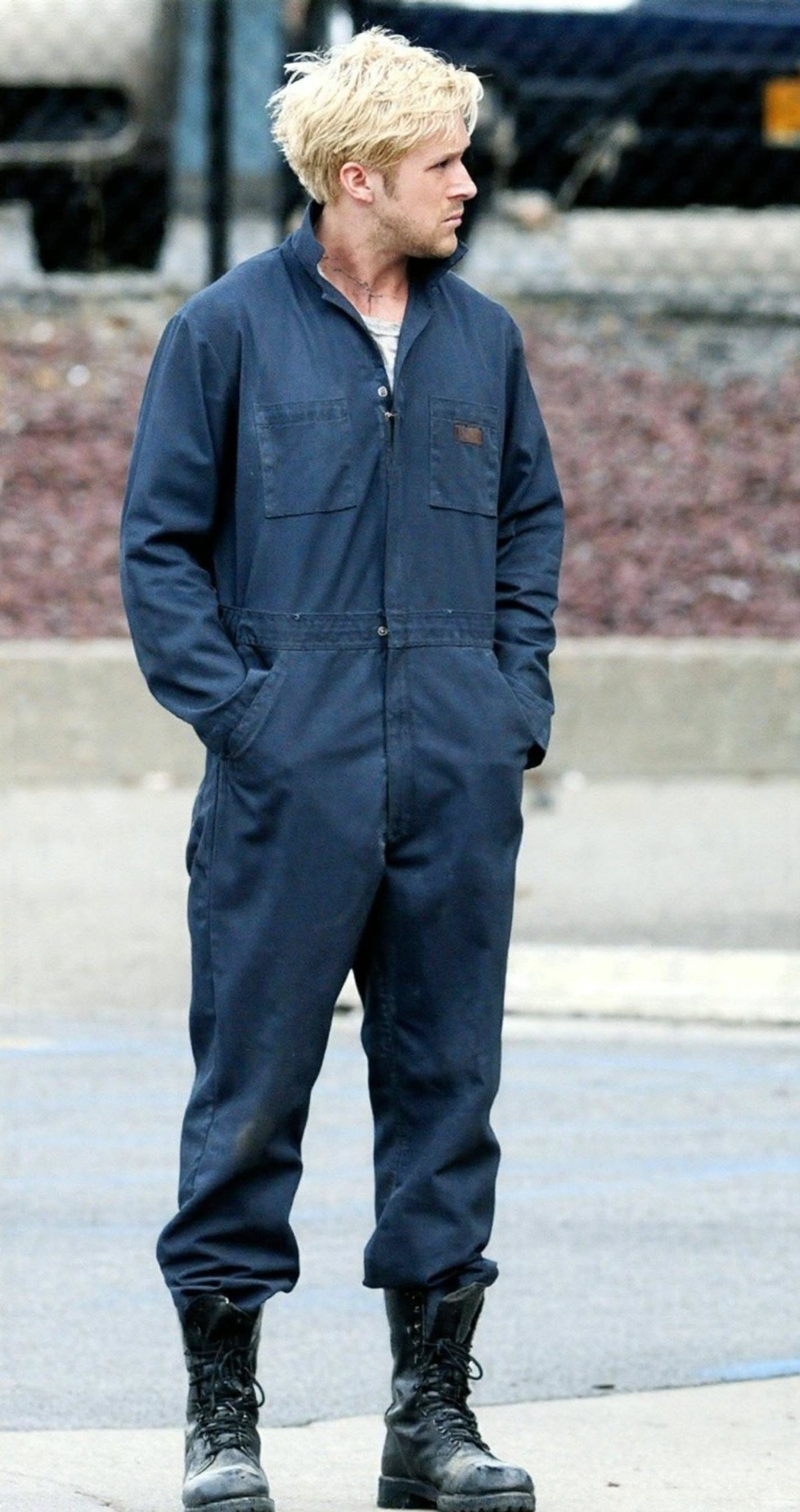 Man with blonde hair wearing navy blue coveralls and black combat boots outdoors.
