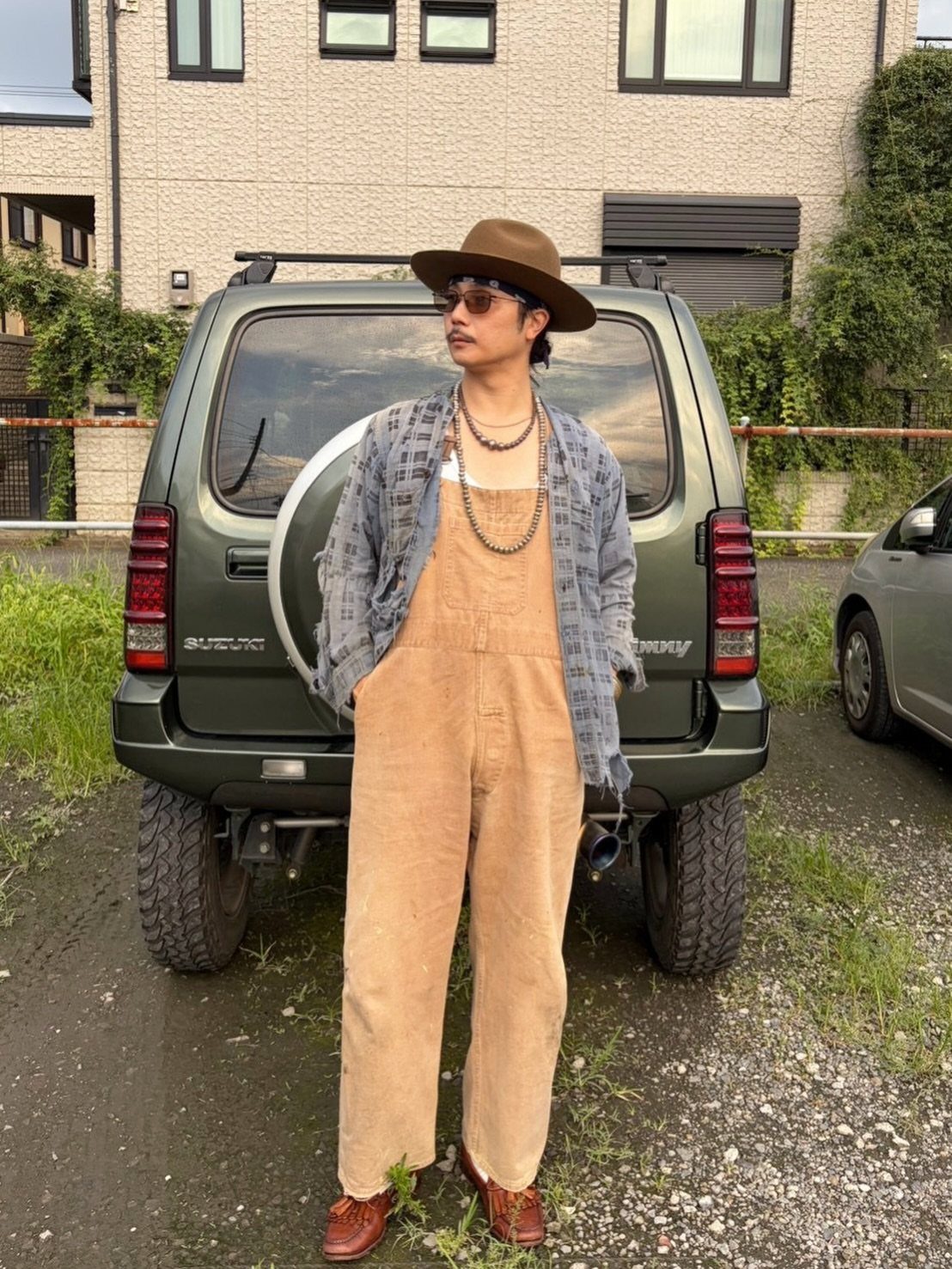 Man in tan corduroy overalls and checkered shirt standing before a green SUV.