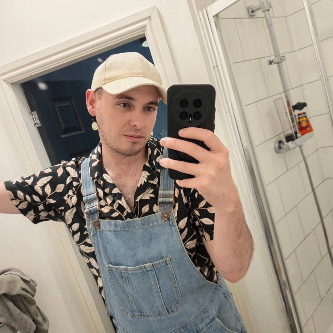Man wearing a floral button-up shirt under light blue denim overalls and beige cap.