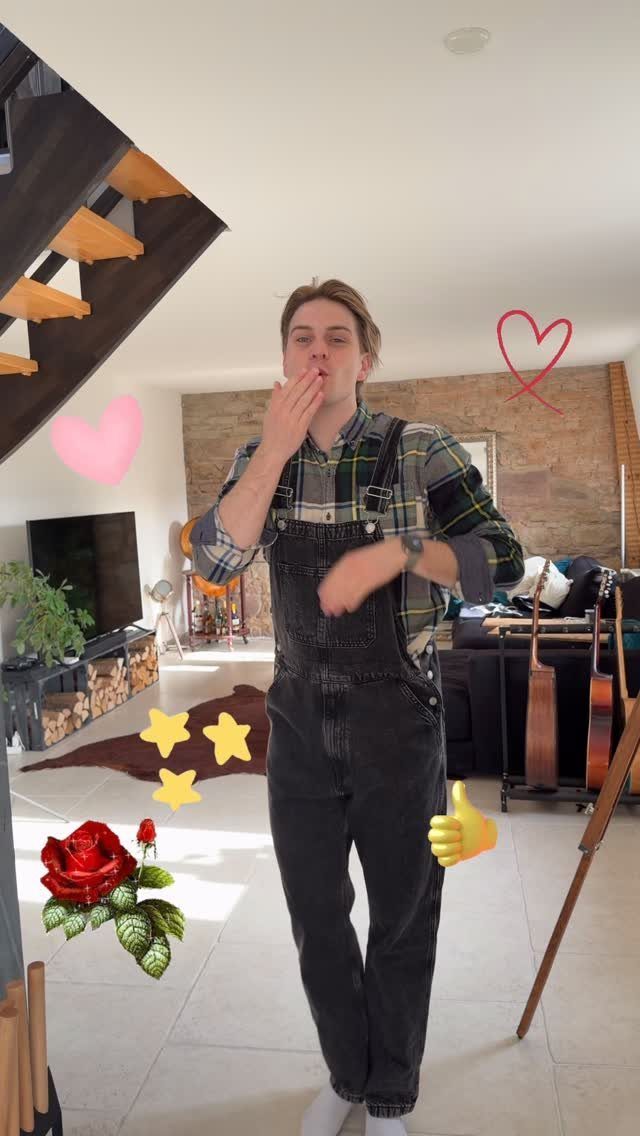 Man wearing a plaid button-up shirt and dark denim overalls in a living room