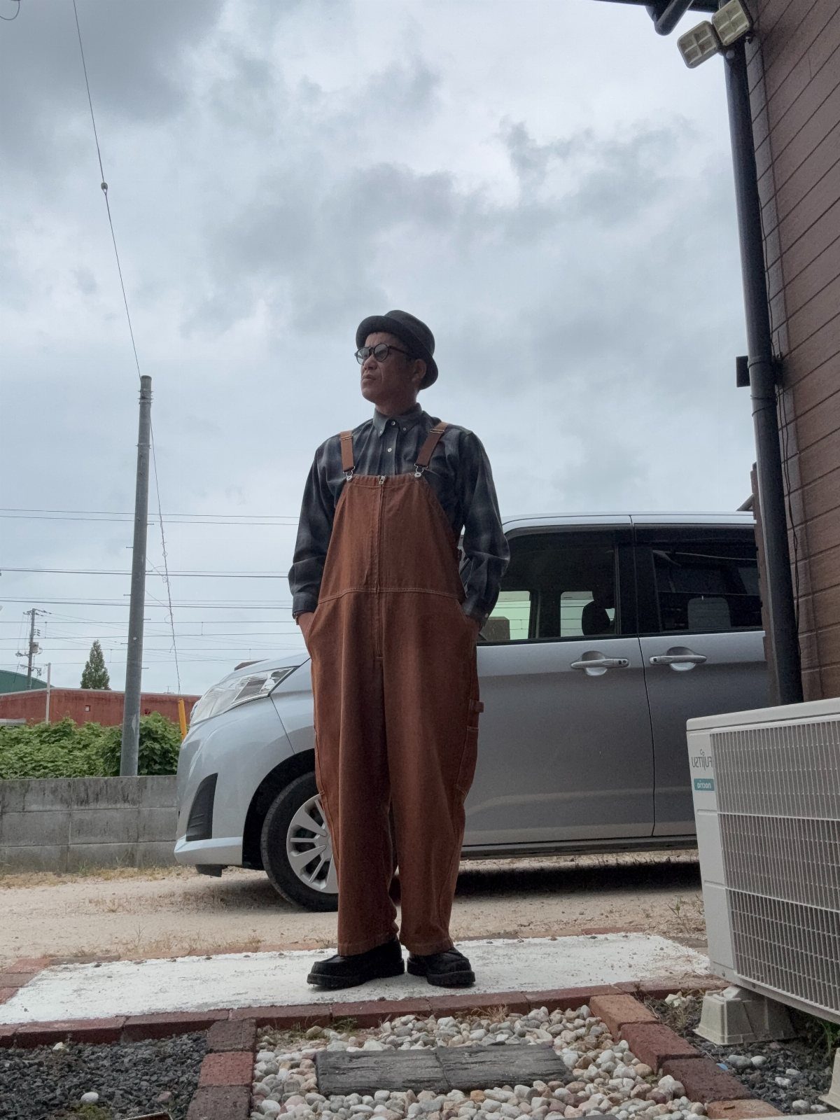 Man wearing brown overalls and a plaid button-up shirt standing outdoors by a car
