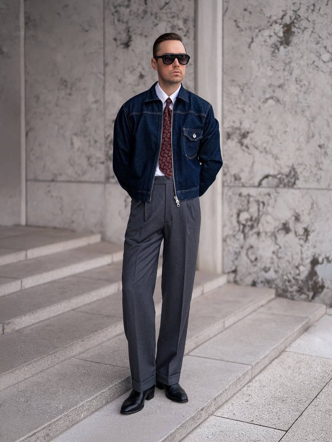 Man wearing a denim jacket and grey tailored trousers standing on stone steps.