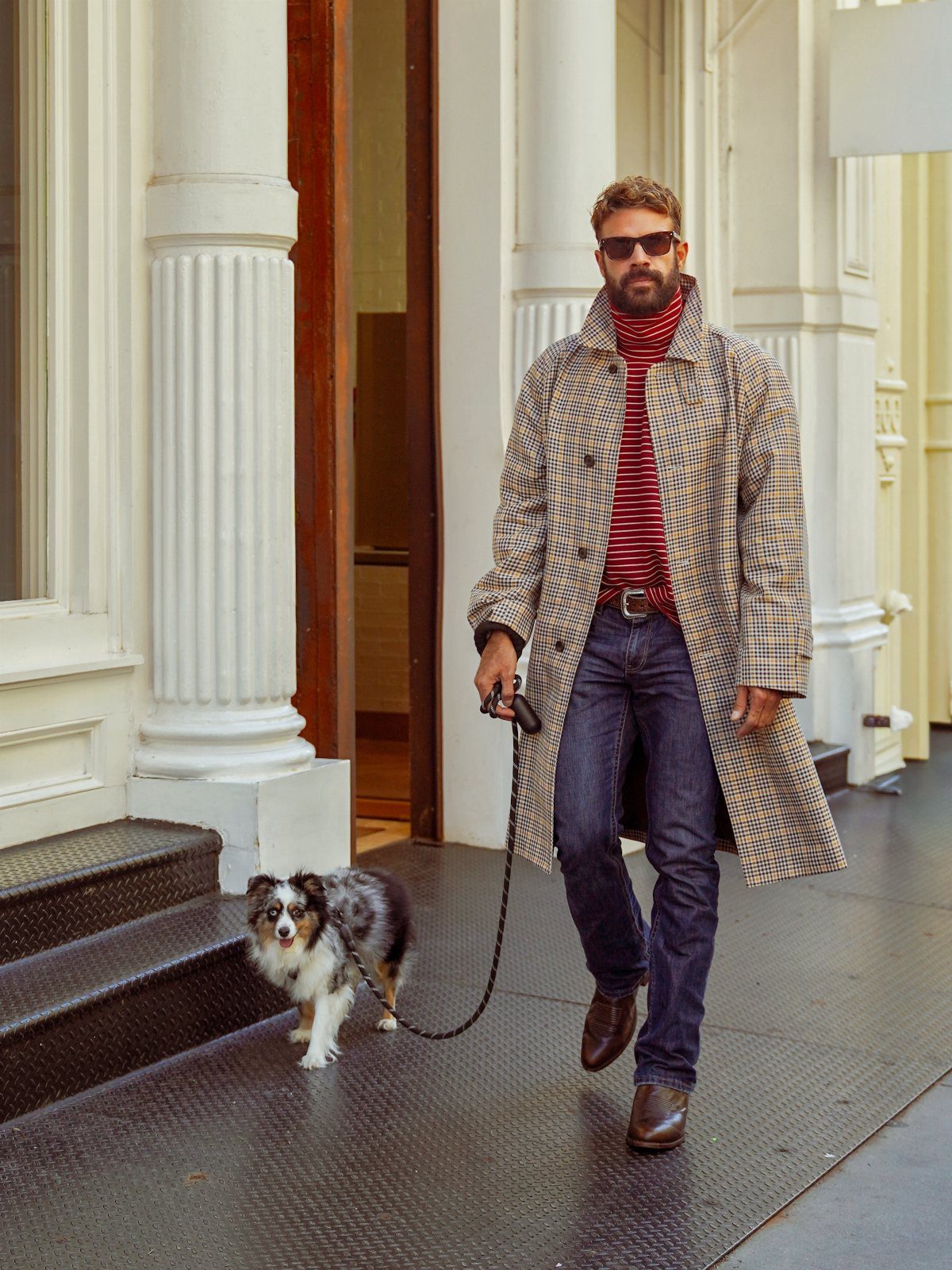 Man in a tan plaid trench coat walking a dog past white columns.