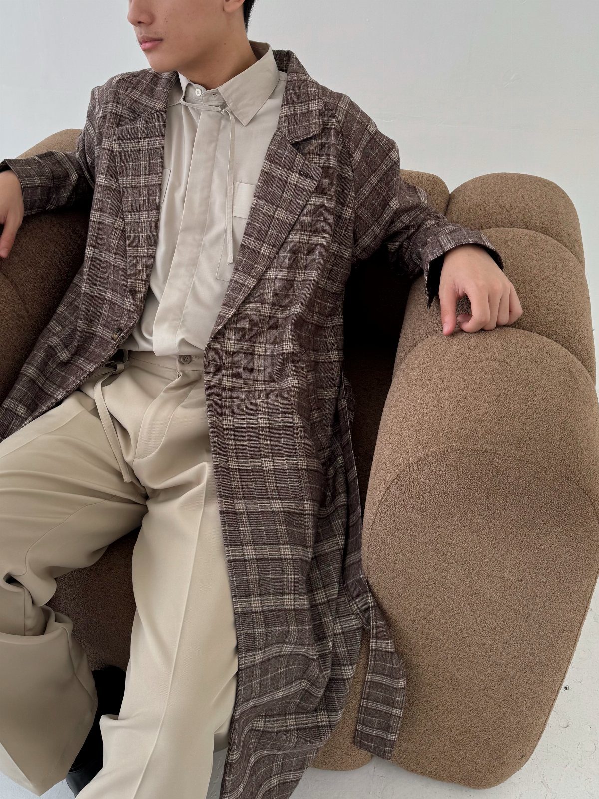 Person wearing a brown plaid trench coat and beige trousers on a curved sofa